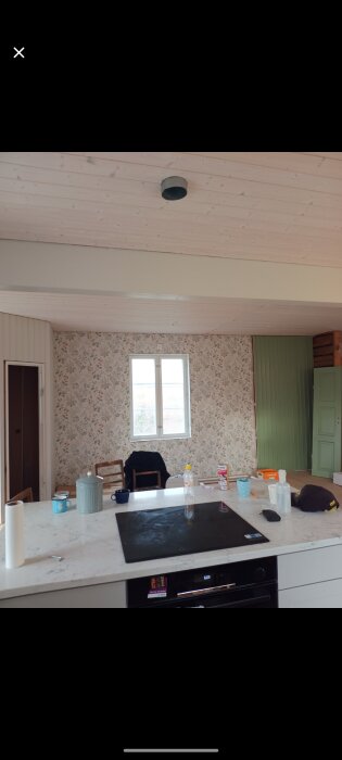 Kök med mönstrad tapet, fönster, gröna dörrar och marmorbänk med köksredskap och en spishäll.