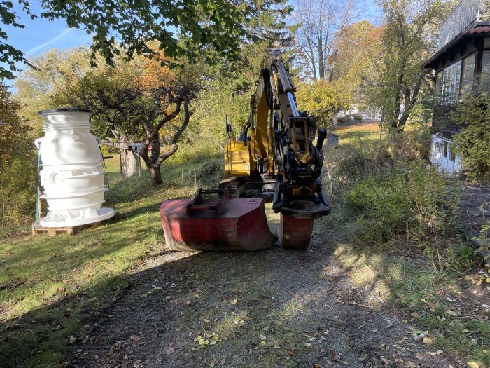 Grävmaskin på en grusväg vid ett hus och en stor vit tank i en trädgård.