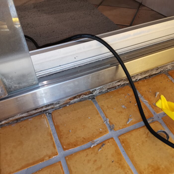Sliten fog vid duschentröskel på kakelgolv, delvis borttagen silikon och träliknande material synligt, svart kabel och gul verktygsspets i bild.