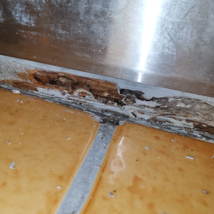 Badrumsgolvet vid dusch med borttagen silikonfog, visar träliknande material och mögelrester på brunorange kakel och metallkanter.