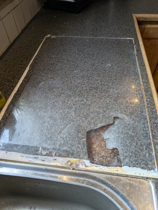 Trasig bänkskiva i ett kök täckt med en plastskiva, synliga skador och del av diskho.