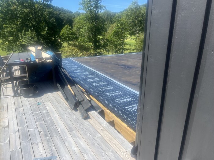 Självhäftande remsor och takmaterial förbereds för att applicera tätskikt på ett tak, med träplattor och skog i bakgrunden.