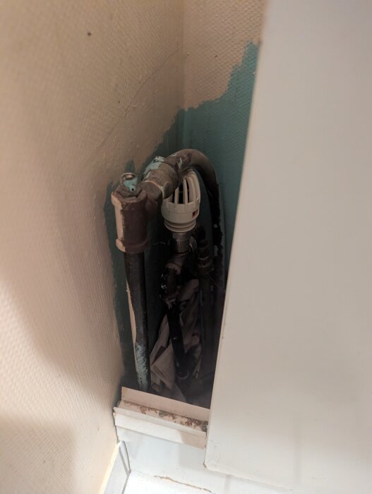 Vägg med exponerade rör och ventil i ett trångt utrymme, omgiven av målad yta och sliten färg.