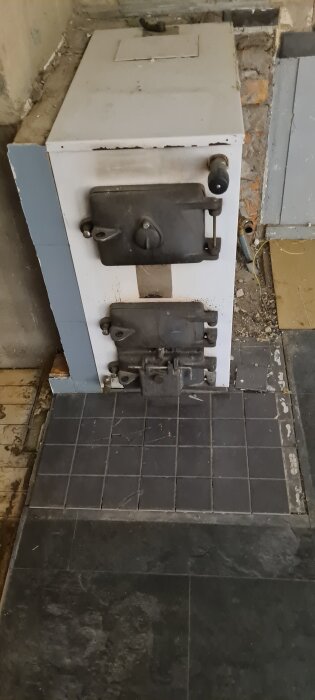 Gammal vedeldad spis med tre luckor, placerad på ett golv täckt med mörka kvadratiska plattor, omgiven av en halvfärdig vägg.