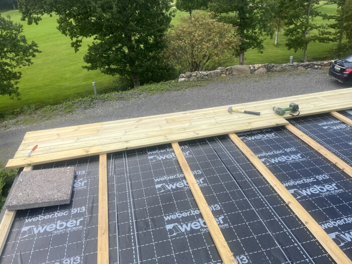 Trallgolv med reglar på webertec 913-täckning, synliga verktyg på plankorna och en vy över grönska och stengärdsgård i bakgrunden.