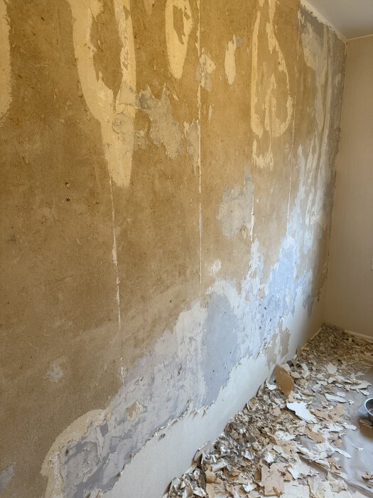 Vägg under renovering med avskrapade tapeter och grått massalager synligt på ytan, rivna tapetbitar på golvet.