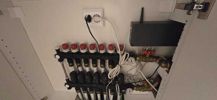 Golvvärmesystem med rör och ventiler, uppkopplat till ett eluttag och en digital enhet, där vissa delar har röda lock.