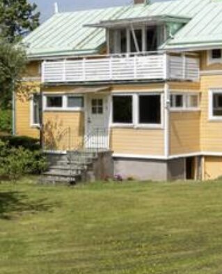 Sekelskifteshus med gul fasad och vit veranda. Grön gräsmatta framför huset med trappa som leder upp till entrén.