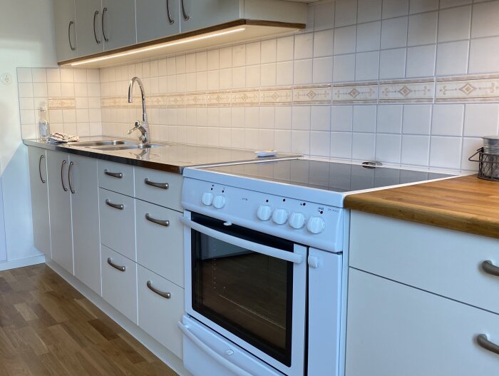 Kök med LED-bänkbelysning, vita kakelplattor, diskho, spis och träbänkskiva. Belysningen sitter monterad nära skåpet.