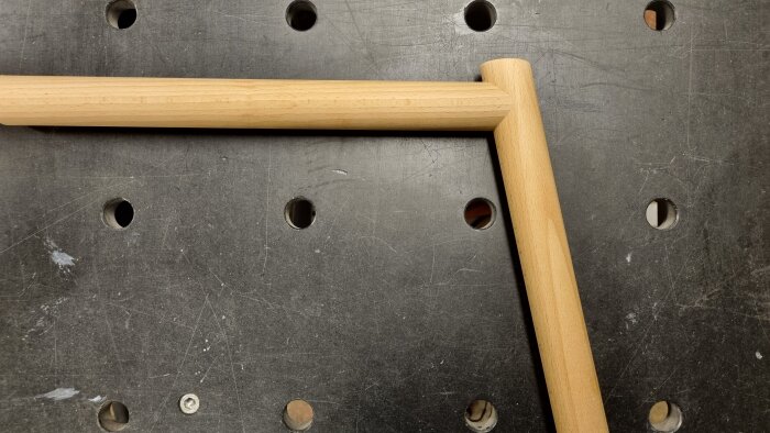 Rund träsammansättning av bok, med två delar i vinkel och samma diameter, på ett arbetsbänk med hål för verktyg.