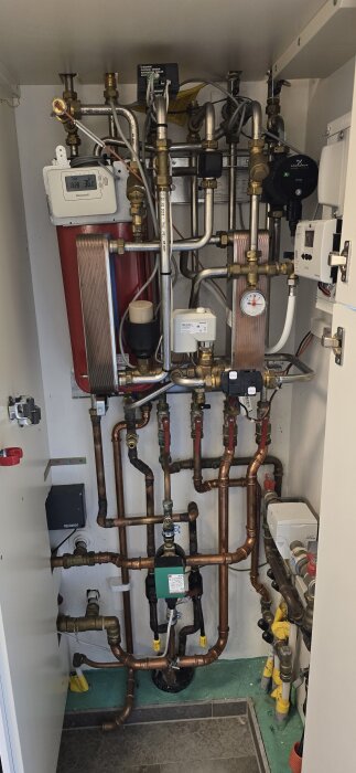 Rörledningar, ventiler och pumpar i ett tekniskt utrymme för VVS. Ett expansionskärl syns till vänster och en termometer mitt i bilden.