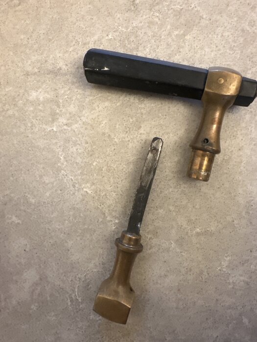Dörrhandtag i delar, en svart och mässingsfärgad del samt en separat mässingsfärgad del med metallstång, ligger på en ljus yta.