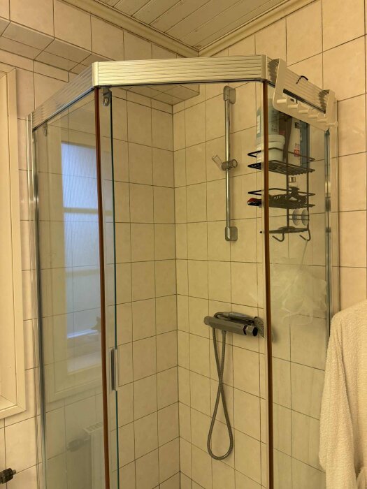 Duschkabin med glasväggar, kromade beslag och en vit kaklad vägg, innehållande en duschslang och hyllor för duschartiklar.