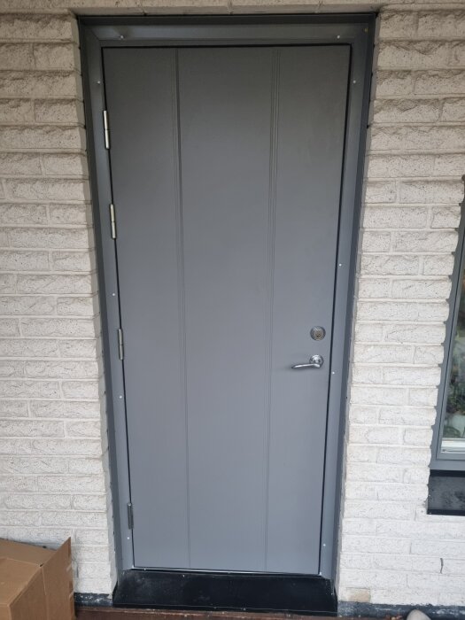 Grå ytterdörr monterad i tegelvägg, sedd utifrån.