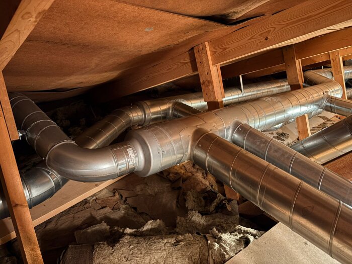 Ventilationsrör under tak i ett vindsutrymme. Rörsystemet är omgivet av träbjälkar och isoleringsmaterial syns på golvet.