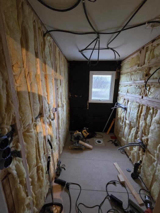 Renovering av toalett med isolering på väggarna, verktyg och sladdar på golvet, samt ett fönster i bakgrunden.