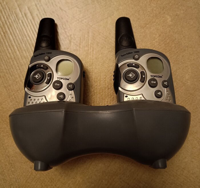 Två gråa walkie-talkies av märket Topcom TwinTalker 1300 i en laddningsstation på ett träbord.