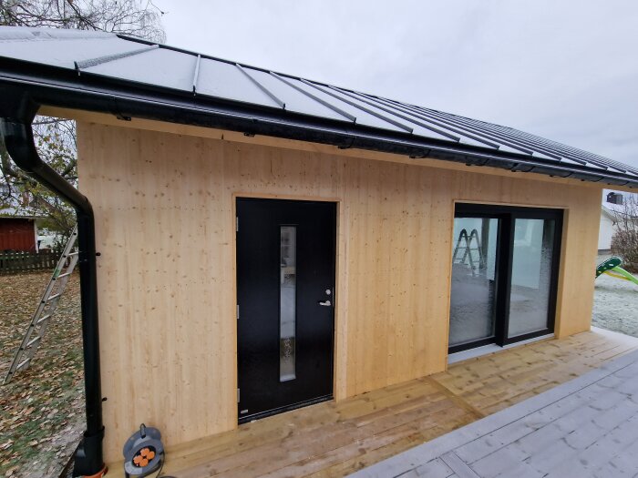 Modernt hus med svart dörr, stora fönster, och svart takränna. Omgivningen är höstig med vissna löv och en stege syns vid sidan.