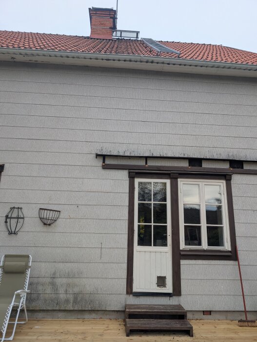 Eternitfasad på hus med röd tegeltak, vit dörr och fönster ovan trädäck. Stege monterad ovanför intill skorstenen på taket.