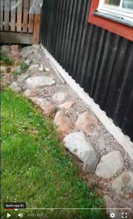 Kant av natursten längs huset med grus och makadam vid sidan av gräsmattan, nära ett svartmålat träpanelhus.