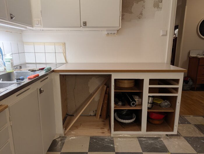 Kök under renovering med målade skåp och öppna hyllor, ofärdig vägg ovanför diskbänken, och ojämnhet där ett tidigare skåp stod.