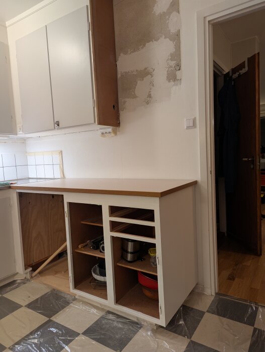 Kök med nybyggt skåp, synliga passbitar och ojämn vägg efter borttaget skåp. Väggen målad, plastskydd på golvet och skåpen delvis öppna.