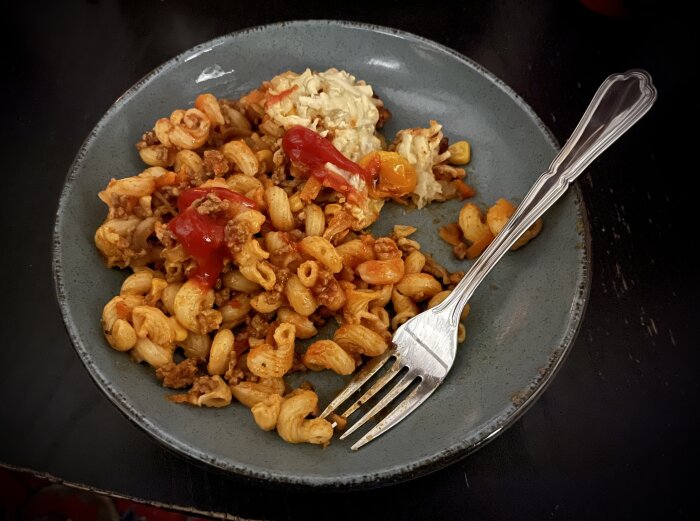 Pastagratäng på grå tallrik med köttfärs, tomat, ost, kryddor, majs, vitkål, morot, vitlök, paprika, lök och ketchup.