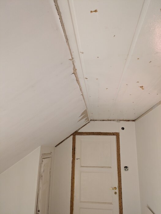 Ojämt avslut mellan gamla och nya gipsskivor i ett snedtak ovanför en dörr i ett hus från 1929 under renovering; synliga skruvhål och skador i taket.
