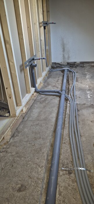 Tätt buntade varmvatten- och kallvattenrör i ett oklart renoverat badrum, med golvrör och träreglar synliga längs en vägg.