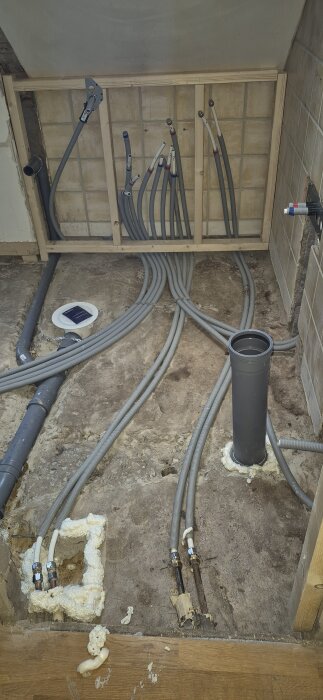 VVS-rör i ett badrum med varm- och kallvattenledningar bundna ihop och golvvärme, omgivna av murade väggar och grovt betonggolv.