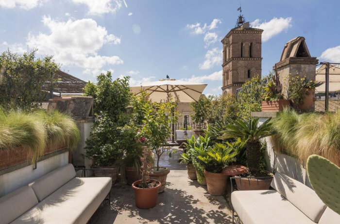 Terrass i Rom med soffor, parasoll, frodig grönska och vy över ett tegelkyrktorn under blå himmel.