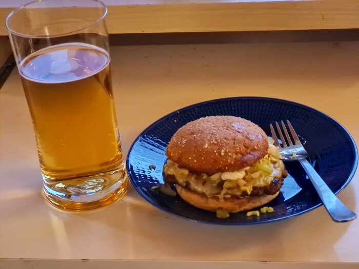 Hamburgare på en blå tallrik med olika ingredienser och ett glas ljus öl på ett bord. Gaffel bredvid hamburgaren.