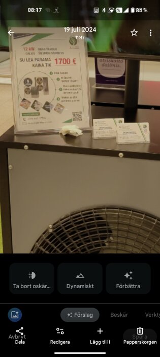 Reklamskylt för Mitsubishi luft vatten, 12 kW, pris 1700 €, på en monter över en fläkt med kontaktuppgifter på papper bredvid.