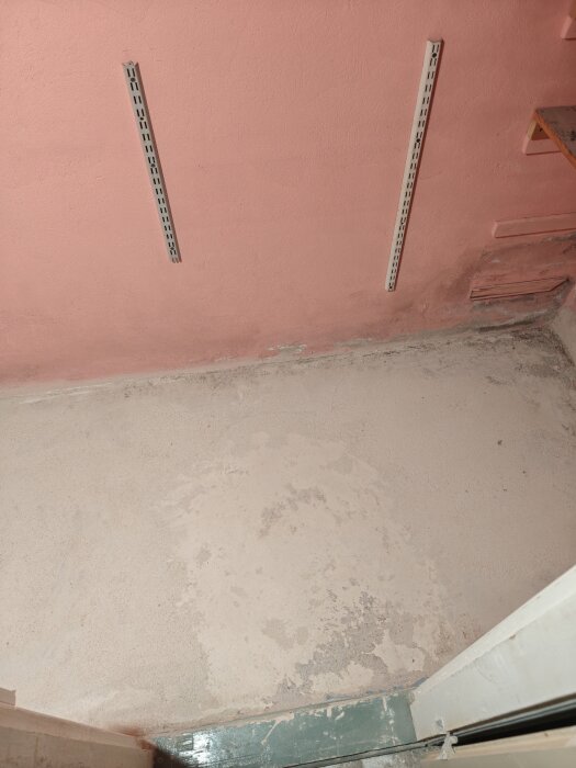 Nyslipat betonggolv i ett källarrum med rosa väggar och metalldetaljer. Tecken på gammal färg och avslitna mattor syns på golvytan.