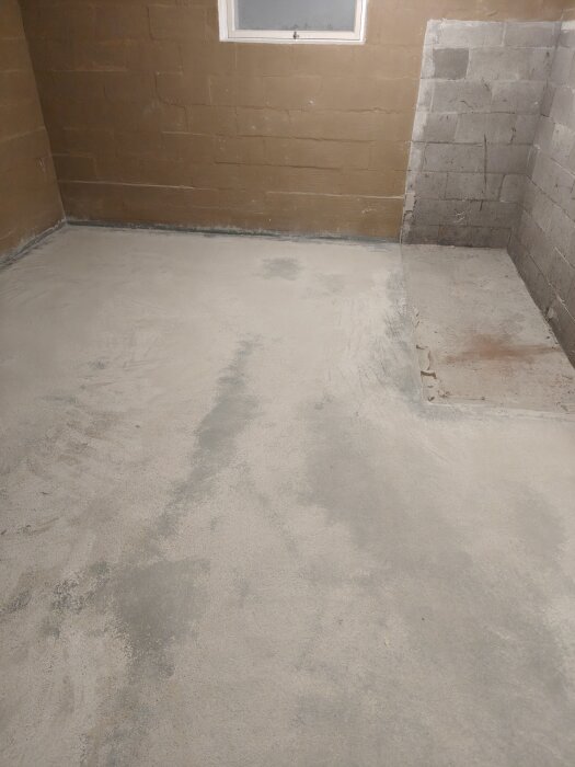 Slipat betonggolv i källare med ojämn yta och en del av väggar i bakgrunden. Ett hörn har en upphöjd sektion mot en blockvägg.