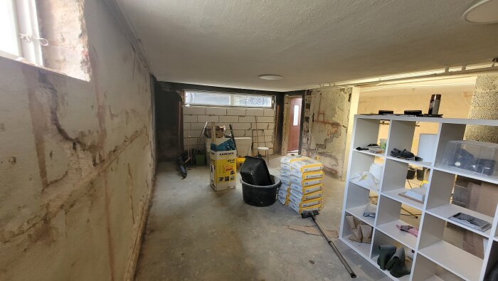 En källare med betongväggar som visar tecken på reparation, verktyg och material står längs väggen, samt en vit hylla fylld med diverse föremål.