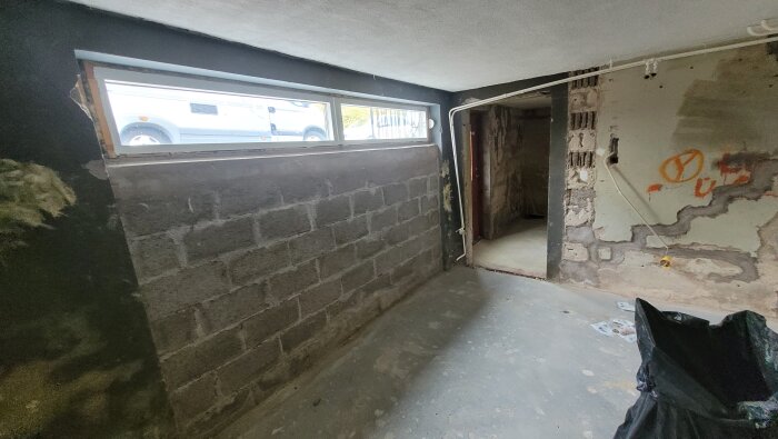 Ojämna betong- och putsytor i ofärdig källare med smalt fönster; synliga mögelspår på väggarna, delvis borttagen puts.