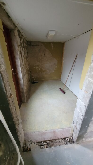 Fuktig källare med avskalat ytskikt och väggar, en sopborste står i hörnet. Lack och puts på väggarna syns delvis borttaget. Gammal dörr till vänster.