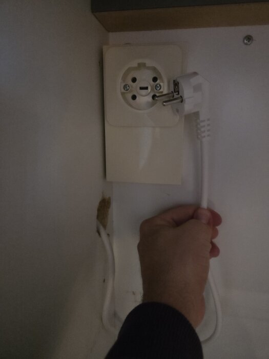 Inifrån ett skåp syns en hand som håller i en vit sladd, ansluten till ett Perilex-uttag.