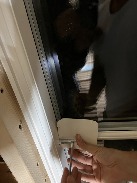 Hand justerar skruv vid ett fönster med delad karm, problem med spröjs och täcklock, i dåligt belyst rum.