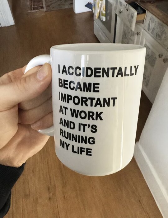 Mugg med text: "I accidentally became important at work and it's ruining my life", hålls framför ett kök.
