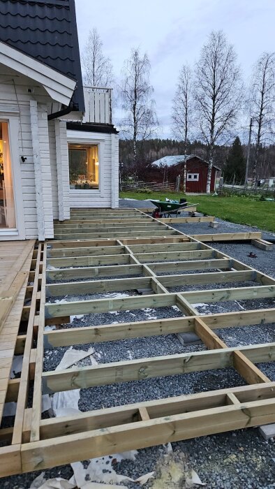 Trädäck under konstruktion, fäst vid ett vitt hus, med träreglar på grusunderlag, och en skottkärra synlig i bakgrunden.