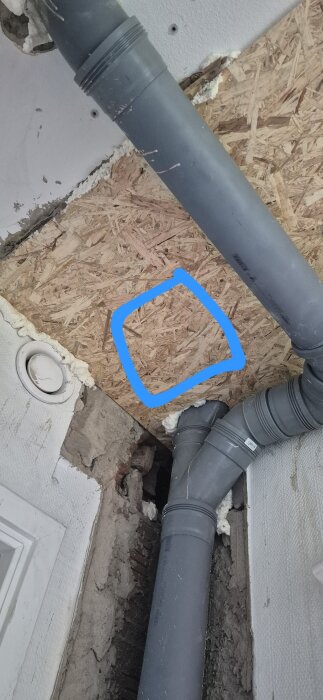 Gråa PVC-rör i en vägg med träpanel, markerad blå ruta där en inspektionslucka ska placeras.