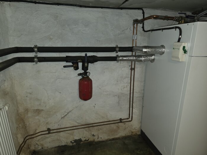 Rostigt expansionskärl på kylrör i källare, kopplat till bergvärmeanläggning. Två VVS-rördelar syns mot en vit vägg.