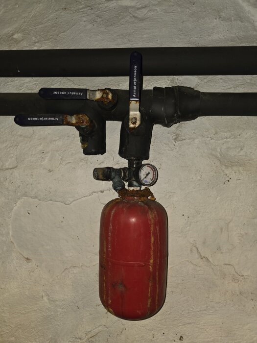 Rostigt expansionskärl i rött med mätare, monterat på ett rörsystem i ett hus med bergvärme.