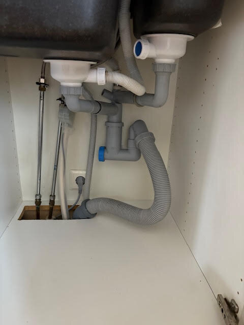 VVS-installation under diskho med grå och vita rör, en blå ventil och en räfflad översvämningsskyddsslang uppåt.