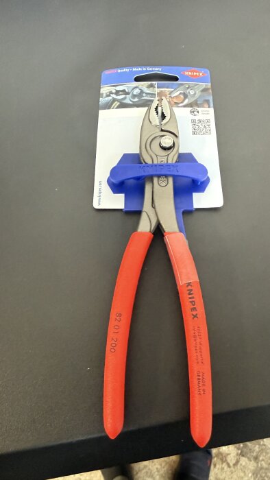 Tång med röda handtag och en blå förpackning från Knipex, placerad på en svart yta.