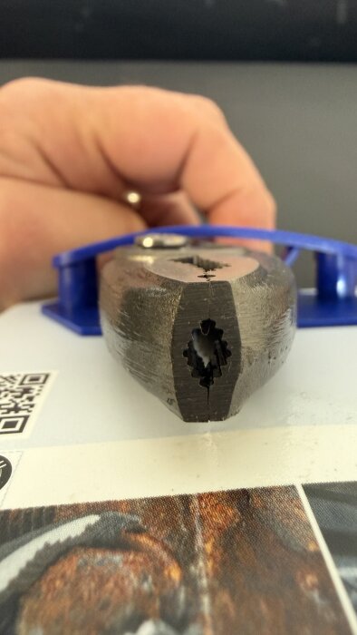 Närbild av en hand som håller en tång nära en blå förpackning, tångens käftar är fokuserade och en QR-kod syns i bakgrunden.