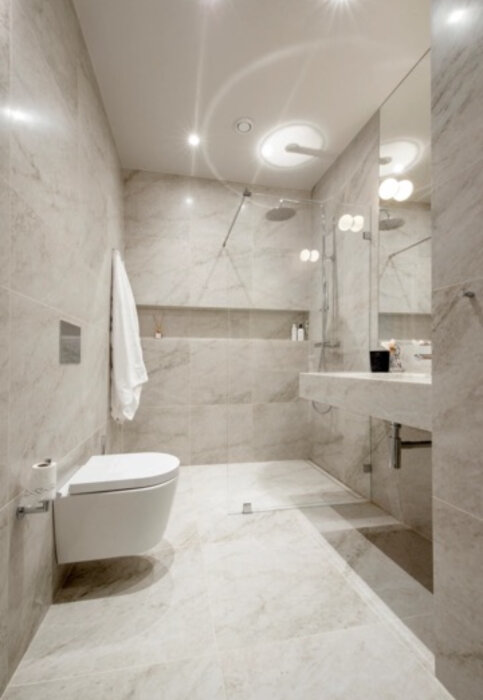 Modernt badrum med Carrara-marmor, vägghängd toalett, dusch med glasvägg, infällda spotlights och stor spegel.