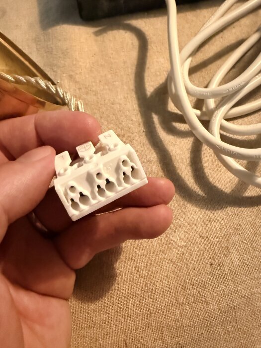 En hand håller en vit elektrisk kopplingsplint med flera hål, bredvid en vit sladd på ett tygbord.